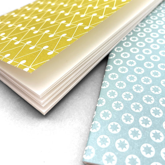 A set of two handcrafted pocket size notebooks by Ola Studio. One cover is a mustard and white triangle geometric design and the other is white stars on an aqua background. Close up of the plain ivory paper