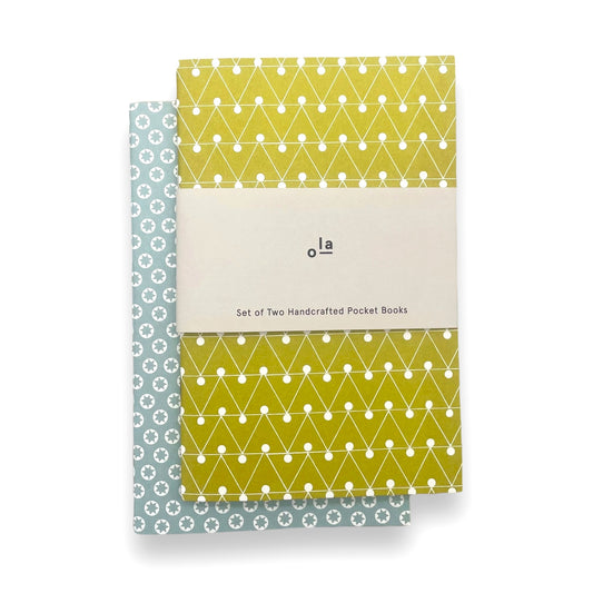 A set of two handcrafted pocket size notebooks by Ola Studio. One cover is a mustard and white triangle geometric design and the other is white stars on an aqua background. Pictured with branded belly band
