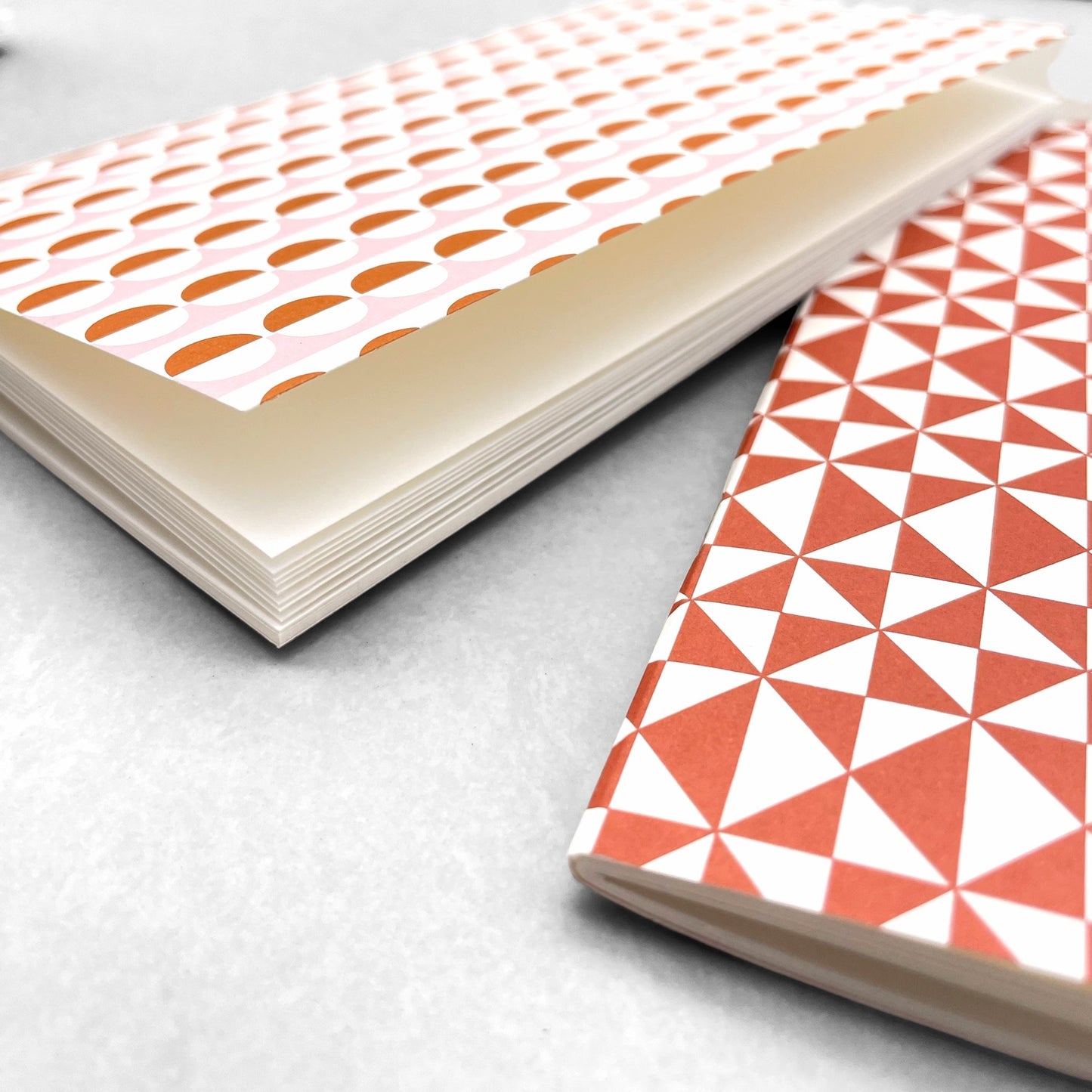 A set of two softcover pocket notebooks by Ola Studio. Two different cover design, one is a circle geometric in pick and orange, the other a triangular geometry in brick red. close up of plain white pages
