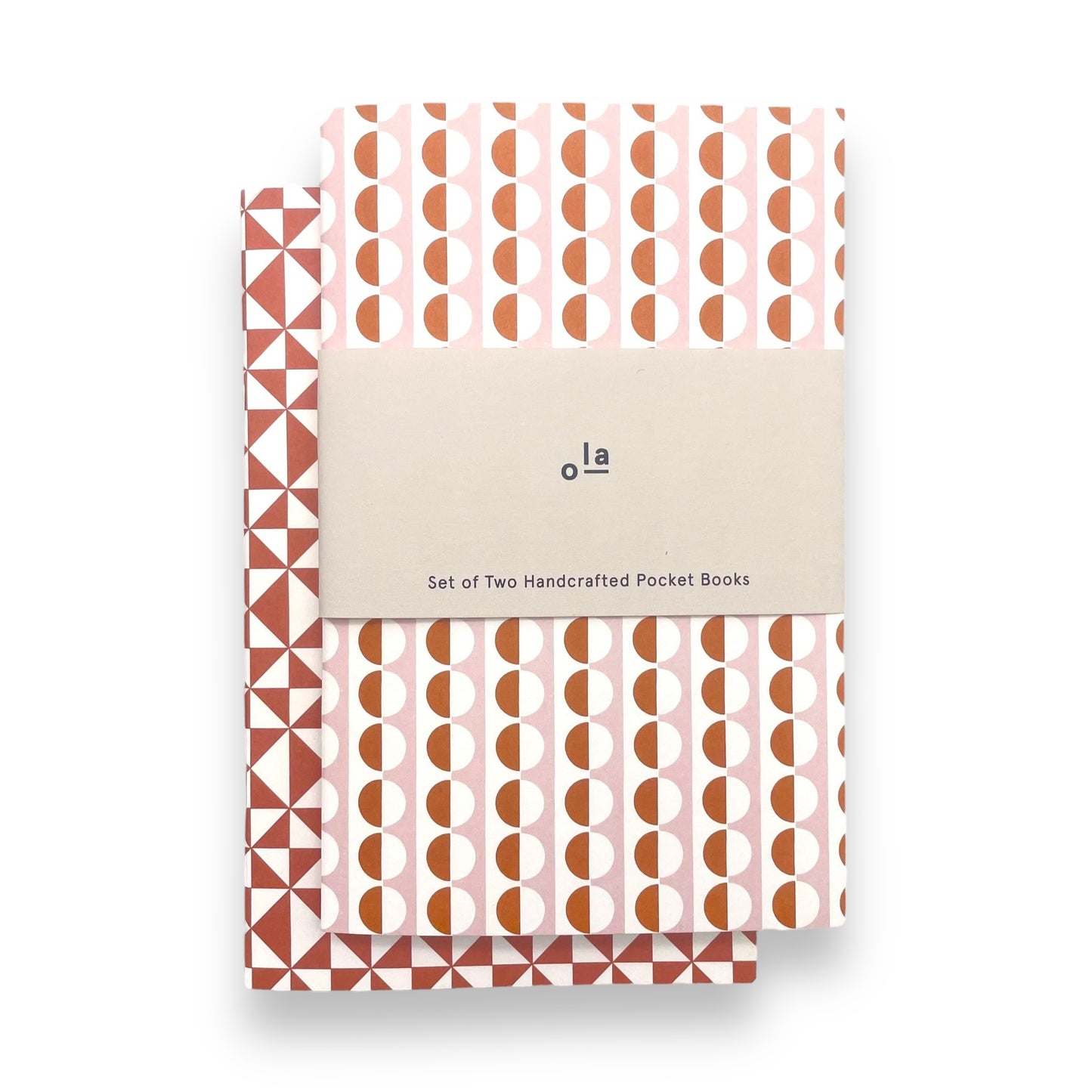 A set of two softcover pocket notebooks by Ola Studio. Two different cover design, one is a circle geometric in pick and orange, the other a triangular geometry in brick red. Pictured with branded band