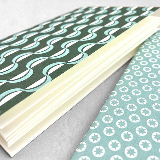 A set of two pocket notebooks by Ola Studio. One cover has a geometric green and aqua wave pattern and the other cover has little white stars on an aqua backdrop. Notebooks have plain pages., close up of the plain ivory pages
