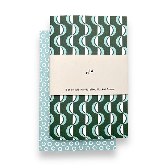 A set of two pocket notebooks by Ola Studio. One cover has a geometric green and aqua wave pattern and the other cover has little white stars on an aqua backdrop. Notebooks have plain pages.