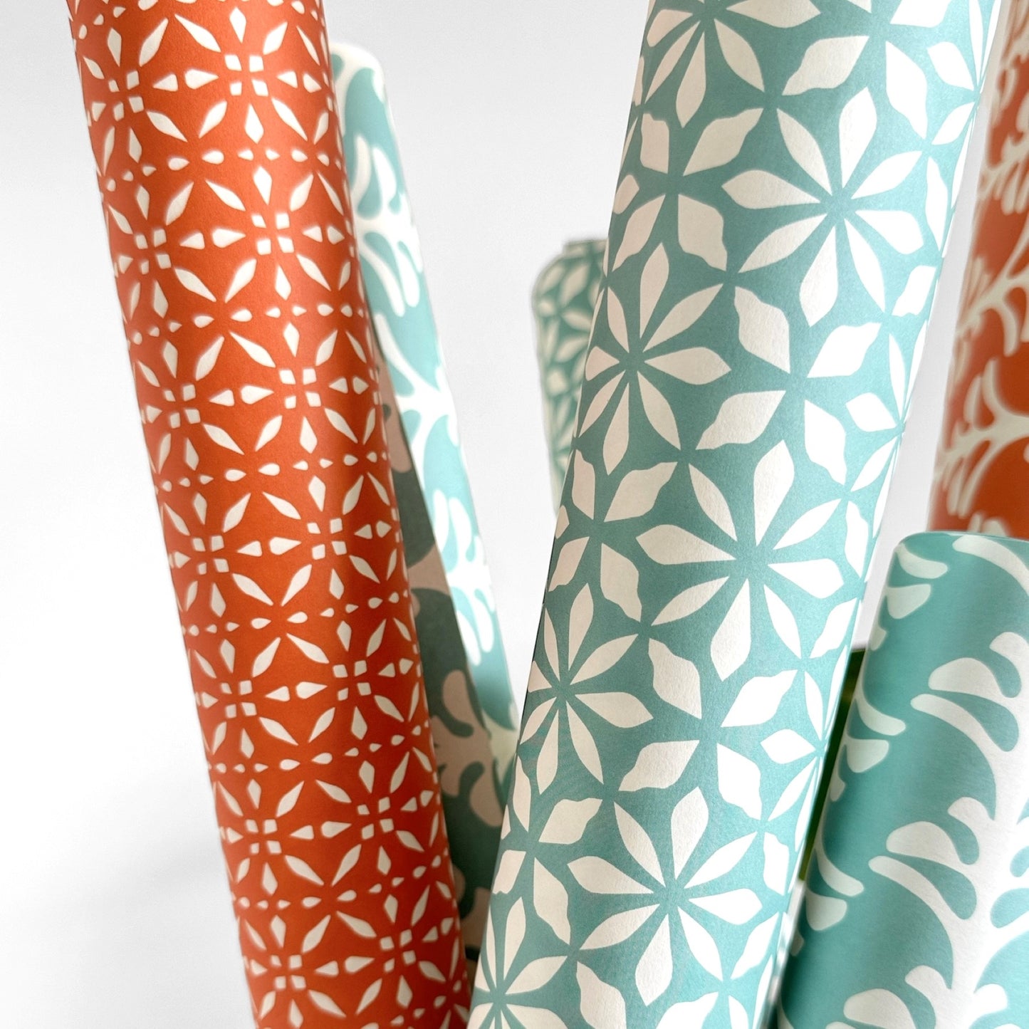 wrapping paper by Otto Editions with a cut-out flower design in white on an aqua blue background. Pictured rolled alongside other designs