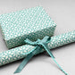 wrapping paper by Otto Editions with a cut-out flower design in white on an aqua blue background. Pictured wrapped as a present with a roll of the paper