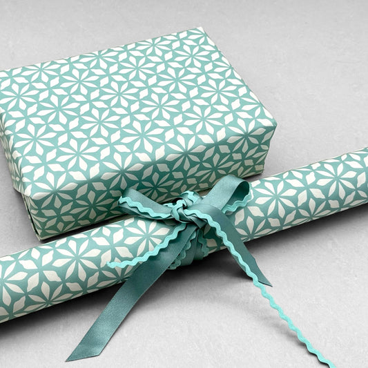 wrapping paper by Otto Editions with a cut-out flower design in white on an aqua blue background. Pictured wrapped as a present with a roll of the paper