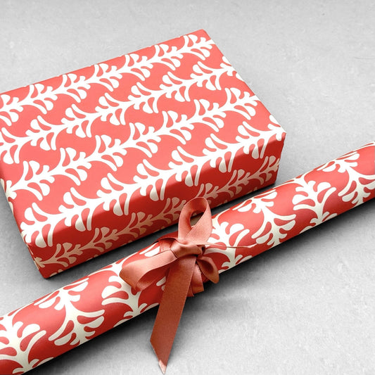 wrapping paper by Otto Editions with a cut-out palm design in white on a coral pink background. Pictured wrapped as a present with a roll of the paper