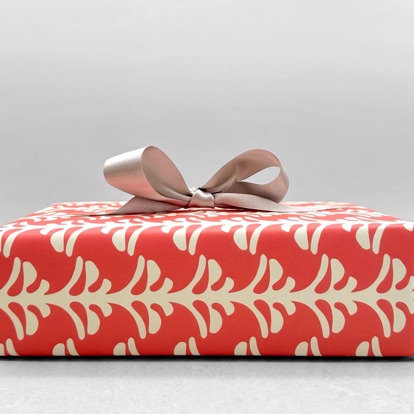 wrapping paper by Otto Editions with a cut-out palm design in white on a coral pink background. Pictured wrapped as a present with a bow