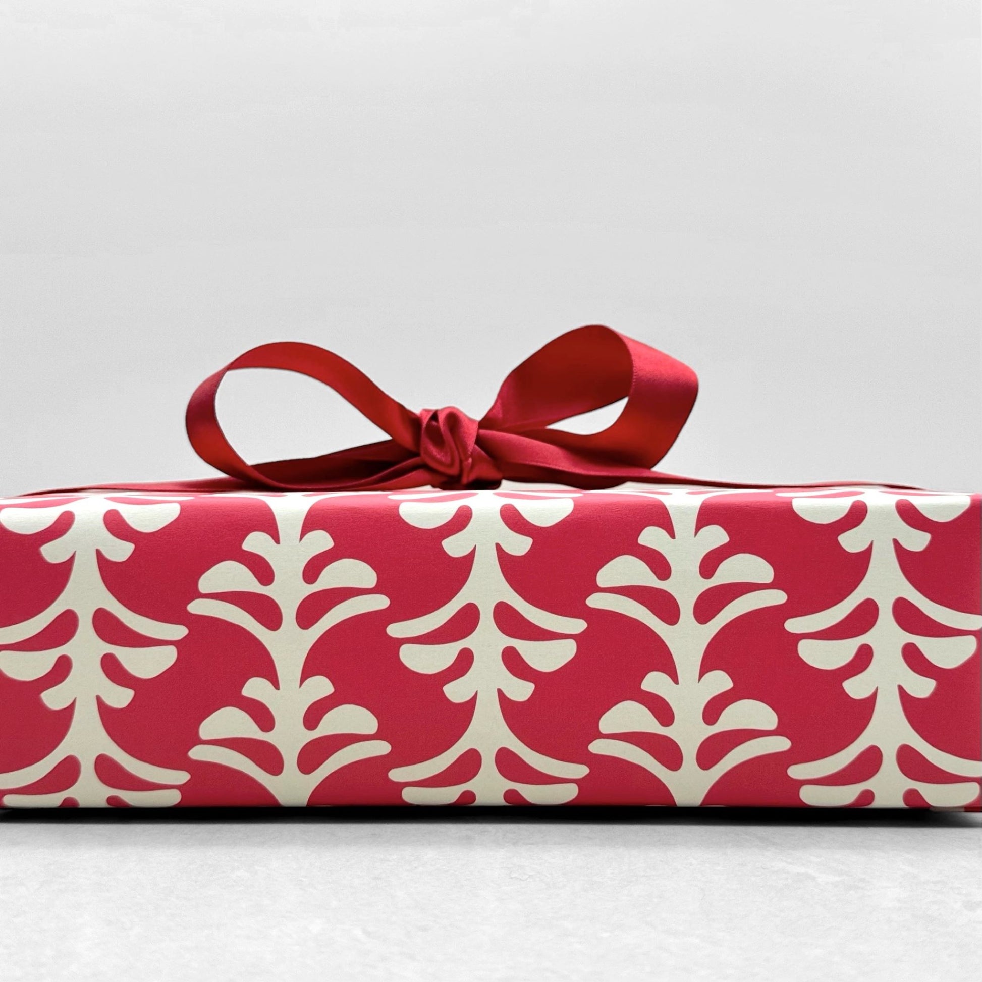 wrapping paper by Otto Editions with a cut-out palm design in white on a hibiscus pink background. Pictured wrapped as a present with a red bow