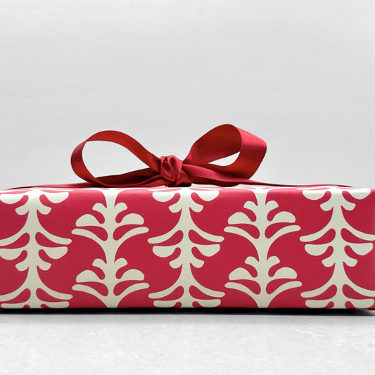 wrapping paper by Otto Editions with a cut-out palm design in white on a hibiscus pink background. Pictured wrapped as a present with a red bow