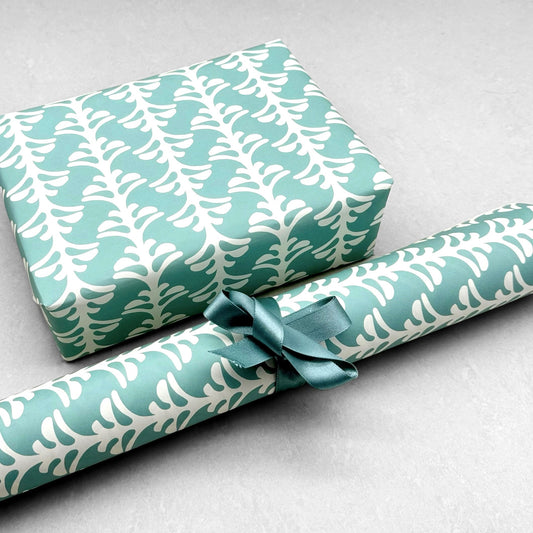 wrapping paper by Otto Editions with a cut-out palm design in white on an aqua blue background. Pictured wrapped as a present with a roll of the paper