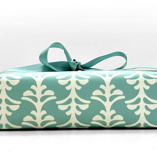 wrapping paper by Otto Editions with a cut-out palm design in white on an aqua blue background. Pictured wrapped as a present with an aqua ribbon