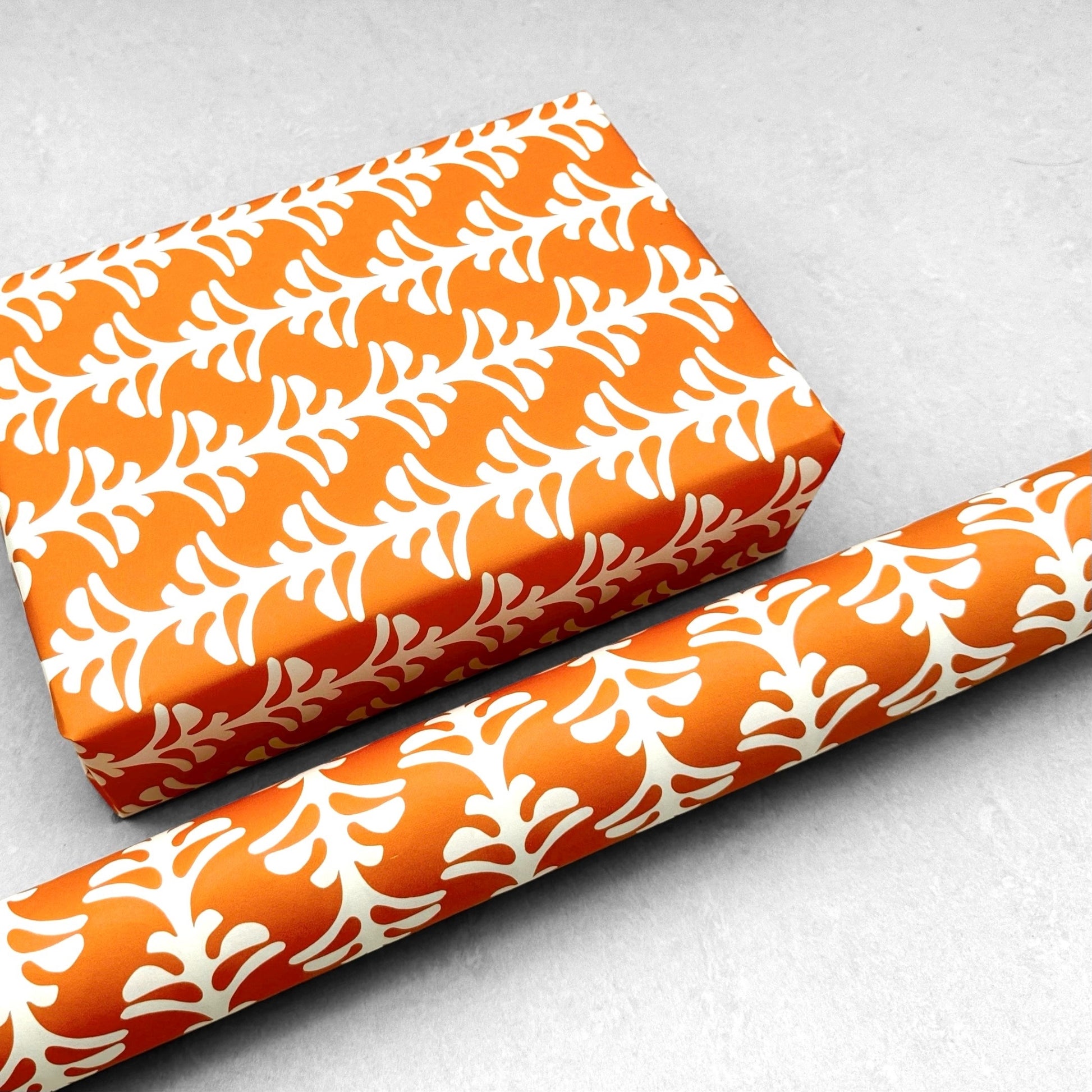 wrapping paper by Otto Editions with a cut-out palm design in white on a spice orange background. Pictured wrapped as a present with a roll of the paper