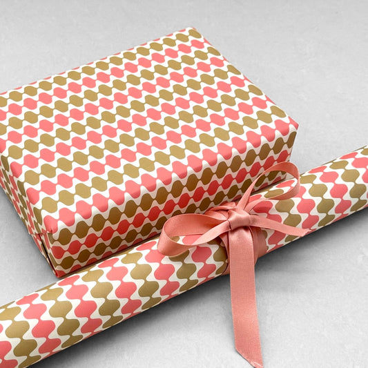 wrapping paper by Otto Editions with a cut-out wavy design in white on a coral and gold background. Pictured wrapped as a present with a roll of the paper
