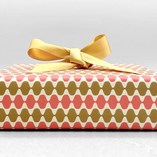 wrapping paper by Otto Editions with a cut-out wavy design in white on a coral and gold background. Pictured wrapped as a present with a gold bow