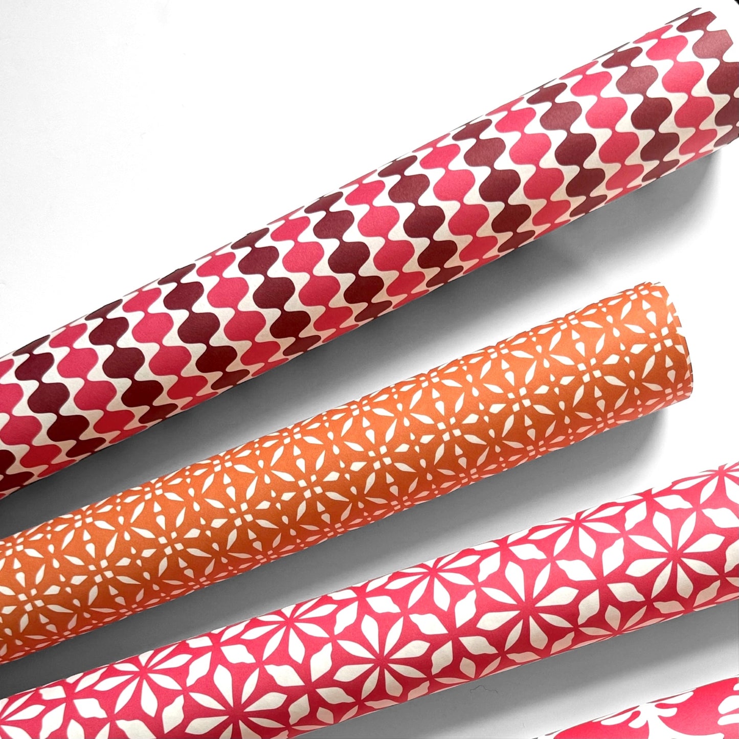 wrapping paper by Otto Editions with a cut-out wavy design in white on a pink and burgundy background. Pictured rolled alongside other designs