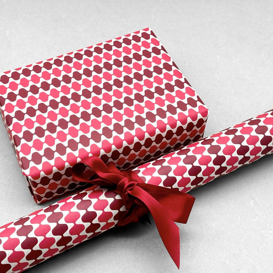 wrapping paper by Otto Editions with a cut-out wavy design in white on a pink and burgundy background. Pictured wrapped as a present with a roll of the paper