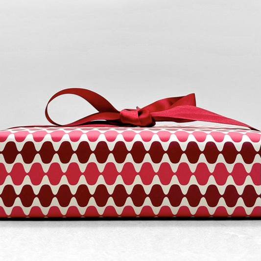 wrapping paper by Otto Editions with a cut-out wavy design in white on a pink and burgundy background. Pictured wrapped as a present with a red bow