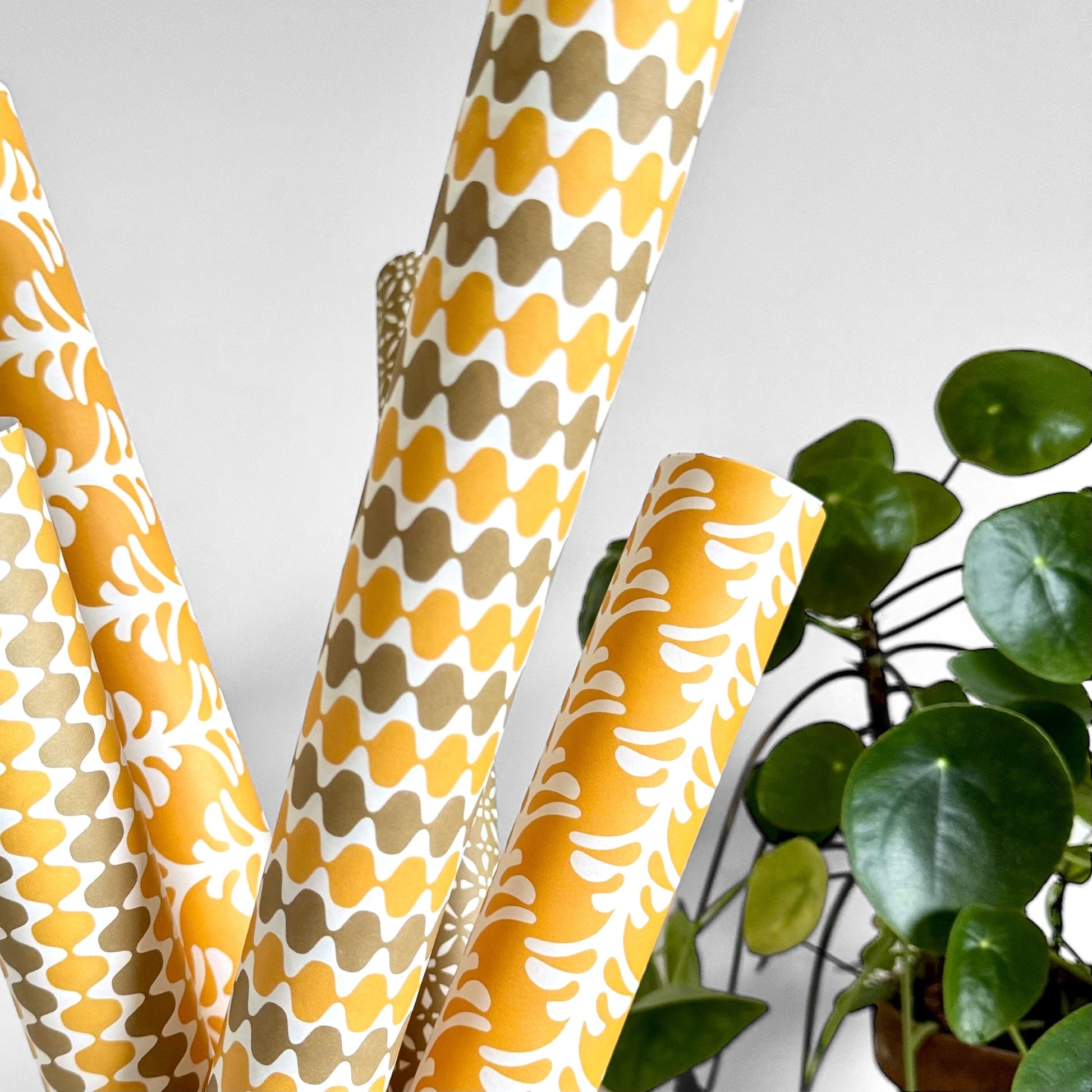 wrapping paper by Otto Editions with a cut-out wavy design in white on a yellow and gold background. Pictured rolled alongside other designs