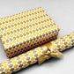 wrapping paper by Otto Editions with a cut-out wavy design in white on a yellow and gold background. Pictured wrapped as a present with a roll of the paper