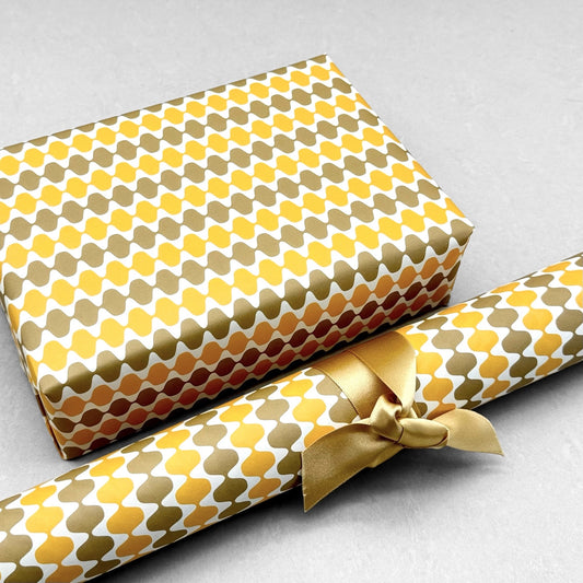 wrapping paper by Otto Editions with a cut-out wavy design in white on a yellow and gold background. Pictured wrapped as a present with a roll of the paper