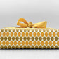 wrapping paper by Otto Editions with a cut-out wavy design in white on a yellow and gold background. Pictured wrapped as a present with a yellow bow