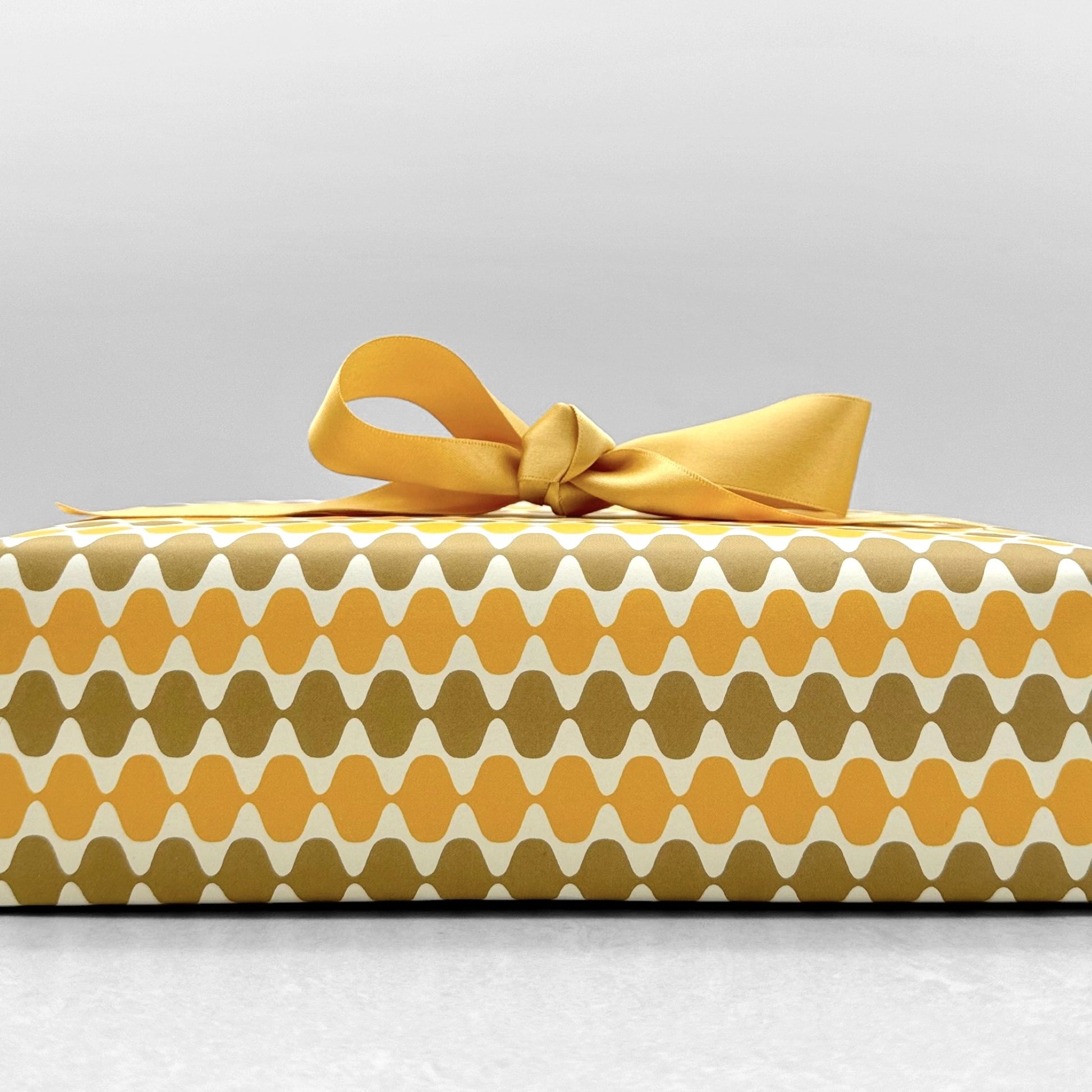 wrapping paper by Otto Editions with a cut-out wavy design in white on a yellow and gold background. Pictured wrapped as a present with a yellow bow