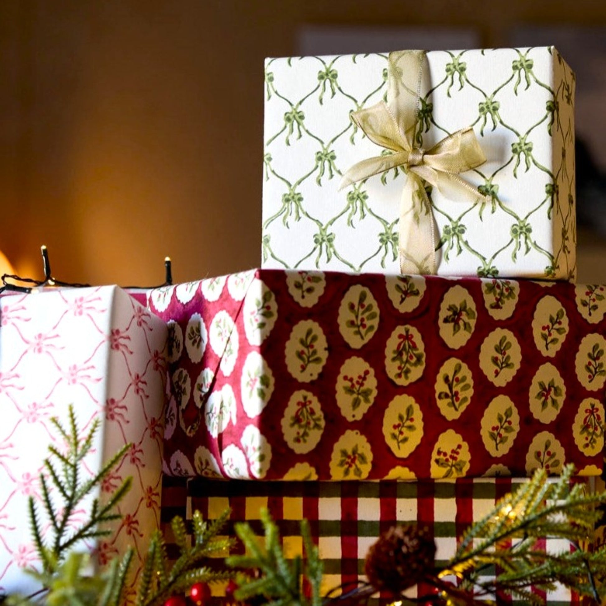 a sheet of hand block printed paper with a pattern of green bows on white. Pictured as a wrapped present with a ribbon at the top of a pile of presents