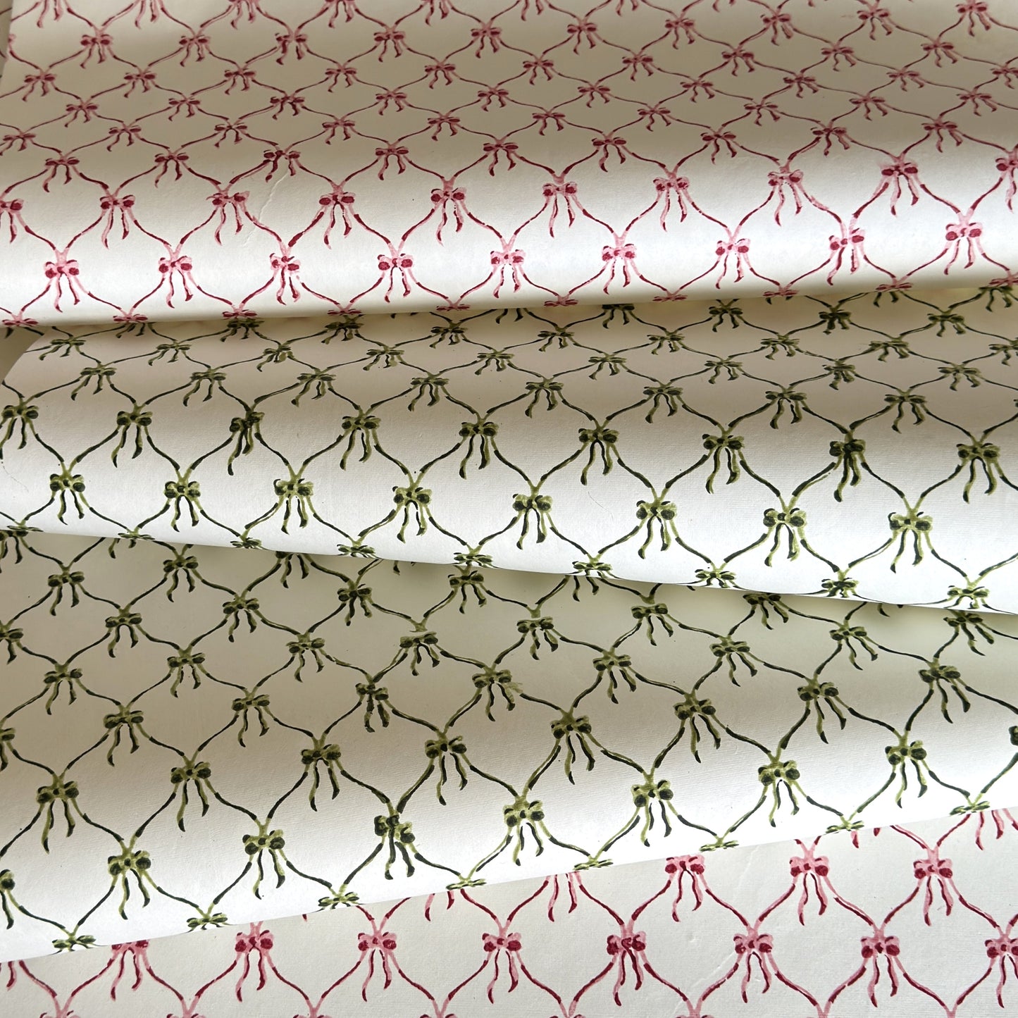 a sheet of hand block printed paper with a pattern of green bows on white, pictured alongside a version in red