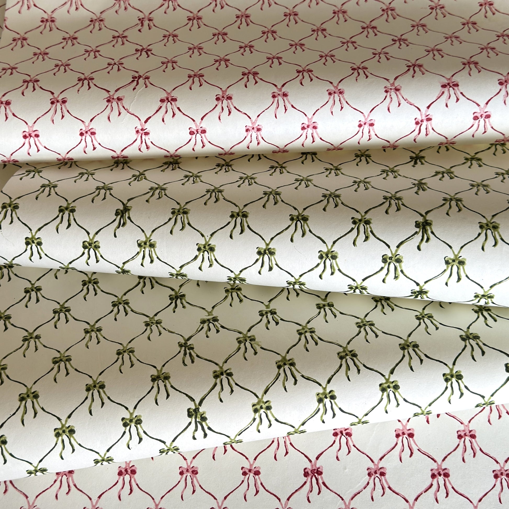 a sheet of hand block printed paper with a pattern of green bows on white, pictured alongside a version in red