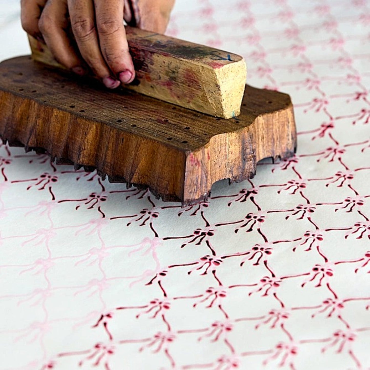 A close-up of a hand holding a wooden printing block over white paper, pressing it to print a repeating red floral pattern. The scene shows the tactile process of textile block printing.
