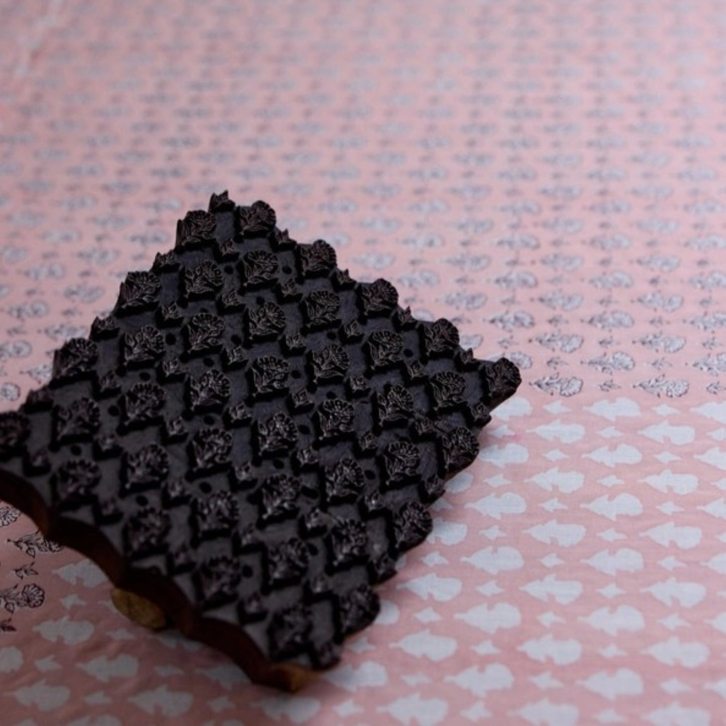 A close-up of a dark brown rectangular stamp or block with a raised floral pattern, placed on a pink background with a repeating light motif.