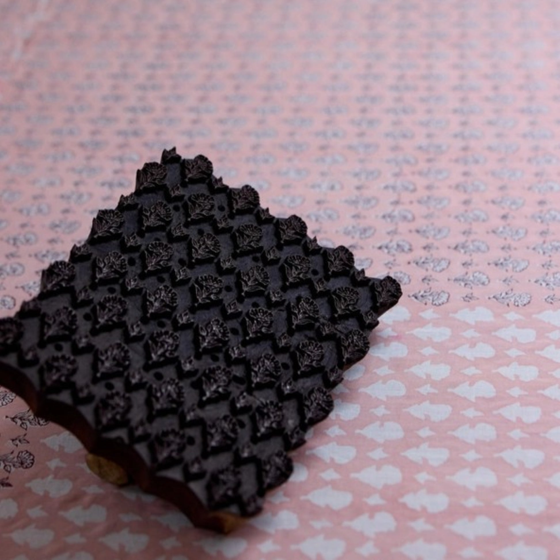 A close-up of a dark brown rectangular stamp or block with a raised floral pattern, placed on a pink background with a repeating light motif.