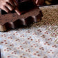 A close-up of a hand pressing a dark wooden block stamp with a scalloped edge onto cotton paper. The pattern on the paper features a blush pink base with an intricate floral and lattice motif in muted sage and dusty rose.