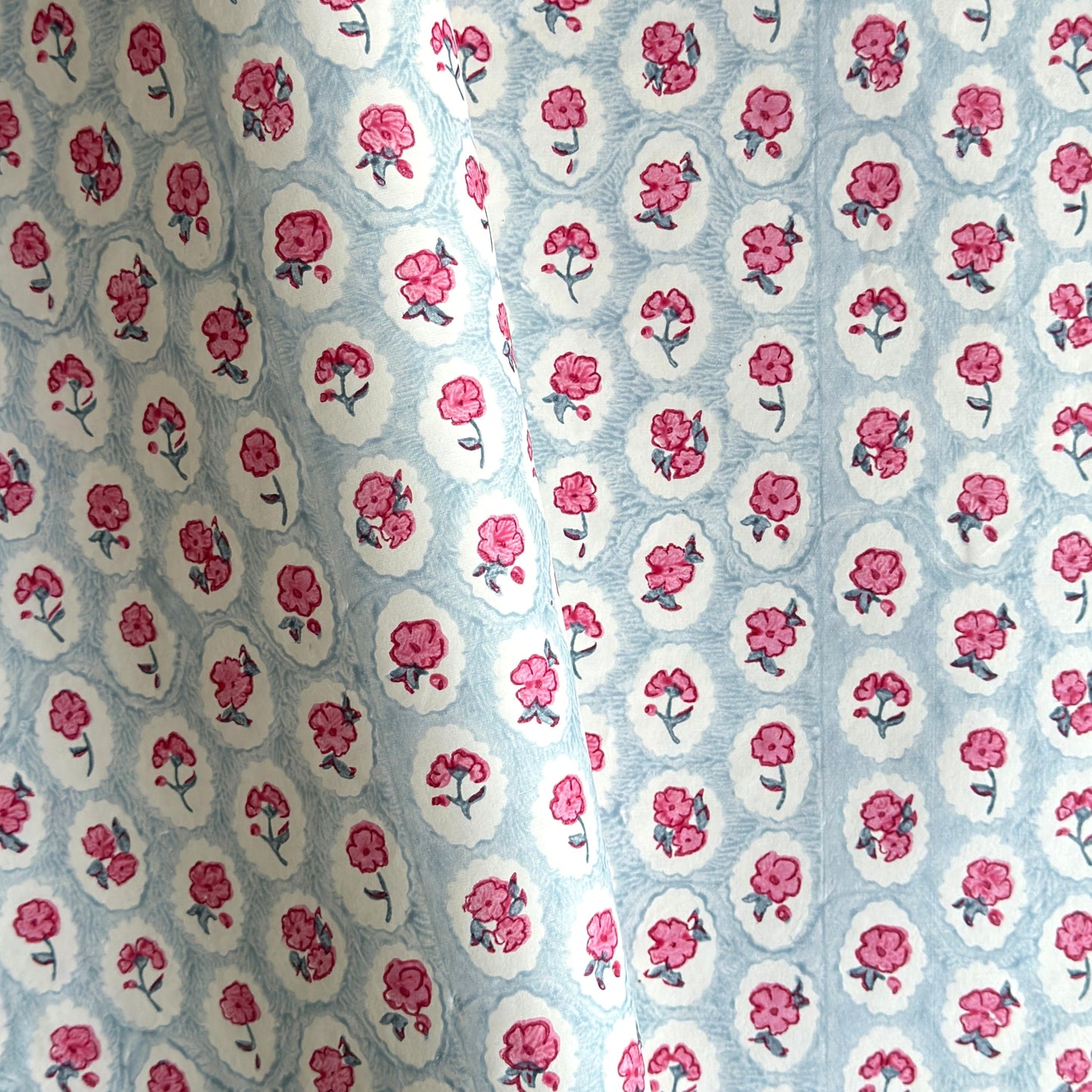 a sheet of hand block printed wrapping paper with a ditsy floral pattern of pink flowers in white ovals with a soft blue backdrop
