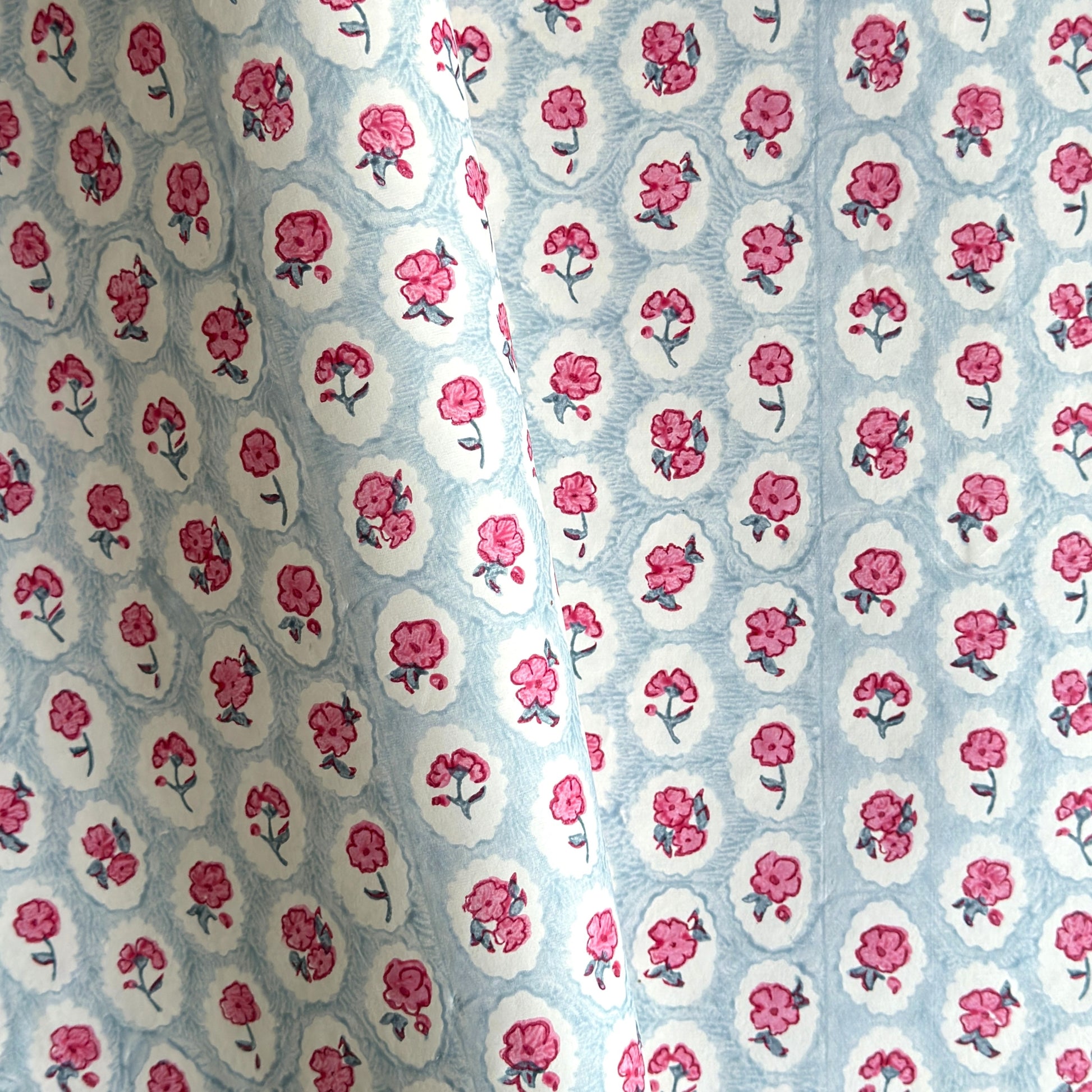 a sheet of hand block printed wrapping paper with a ditsy floral pattern of pink flowers in white ovals with a soft blue backdrop