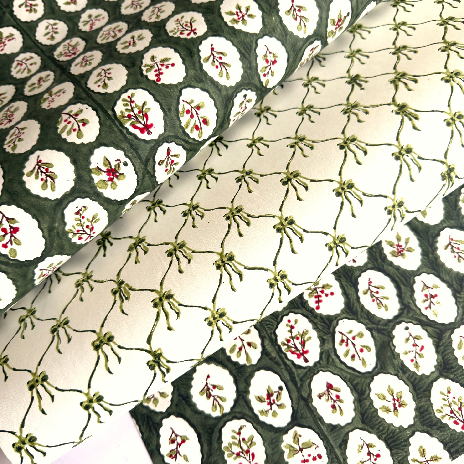 a sheet of block printed paper with a dark green backdrop and white oval scallops with winter foliage motif in the centre.  Pictured with a different design paper with a design of green bows on white