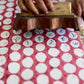 A close-up of a hand holding a wooden hand block printing stamp over white paper with repeated red motifs and black calligraphic marks.