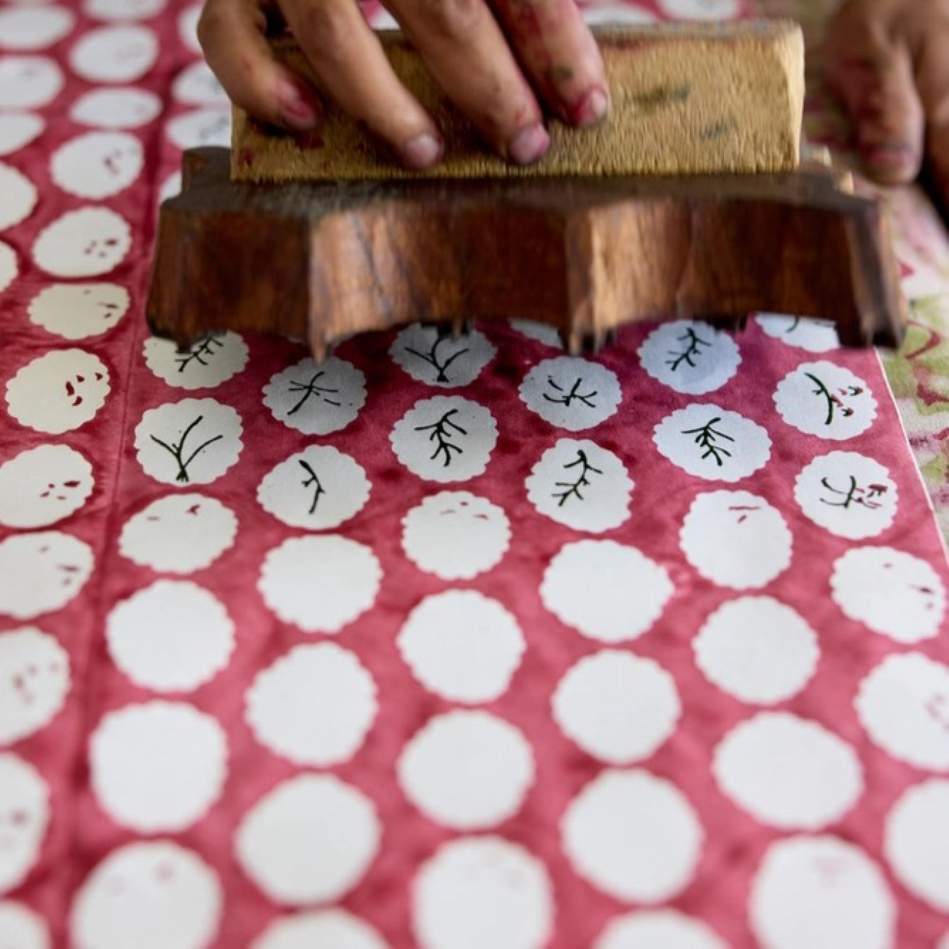 A close-up of a hand holding a wooden hand block printing stamp over white paper with repeated red motifs and black calligraphic marks.