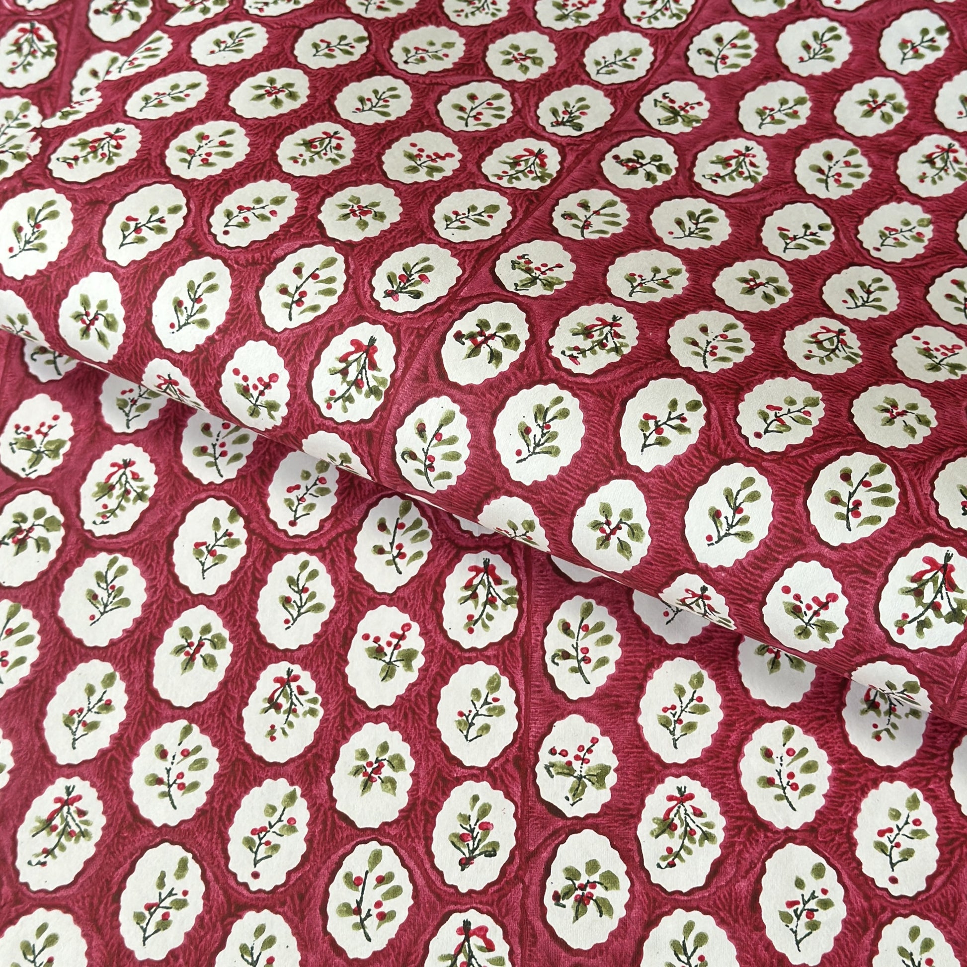 A close-up of a hand block printed paper with a repeating pattern of cream medallions featuring green leaves and red berries on a deep red background.