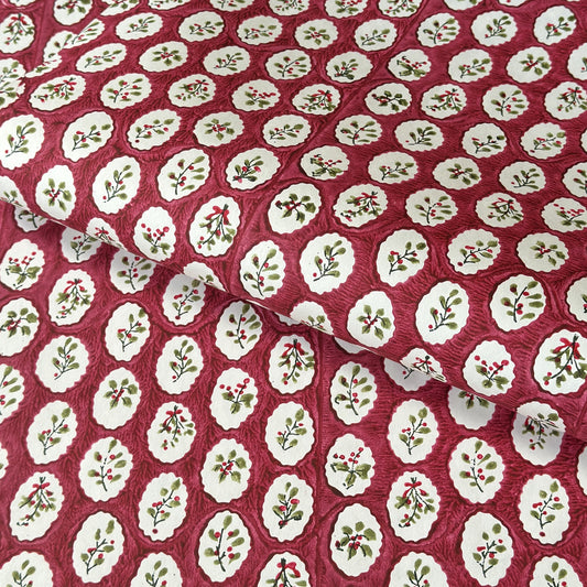 A close-up of a hand block printed paper with a repeating pattern of cream medallions featuring green leaves and red berries on a deep red background.