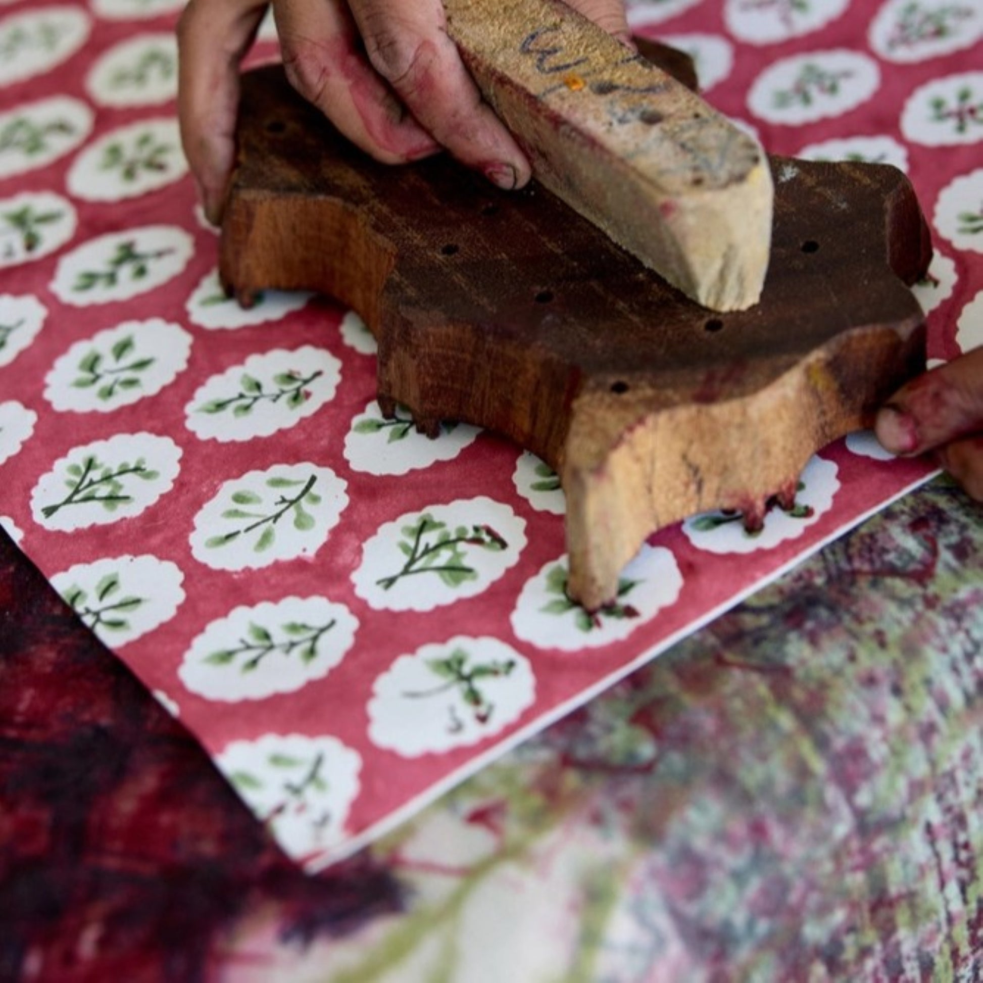A close-up of hands using a wooden stamp with a carved handle to press a red patterned sheet of paper, showing a white floral motif with green leaves.