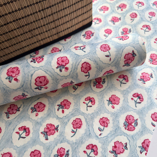 a sheet of hand block printed wrapping paper with a ditsy floral pattern of pink flowers in white ovals with a soft blue backdrop