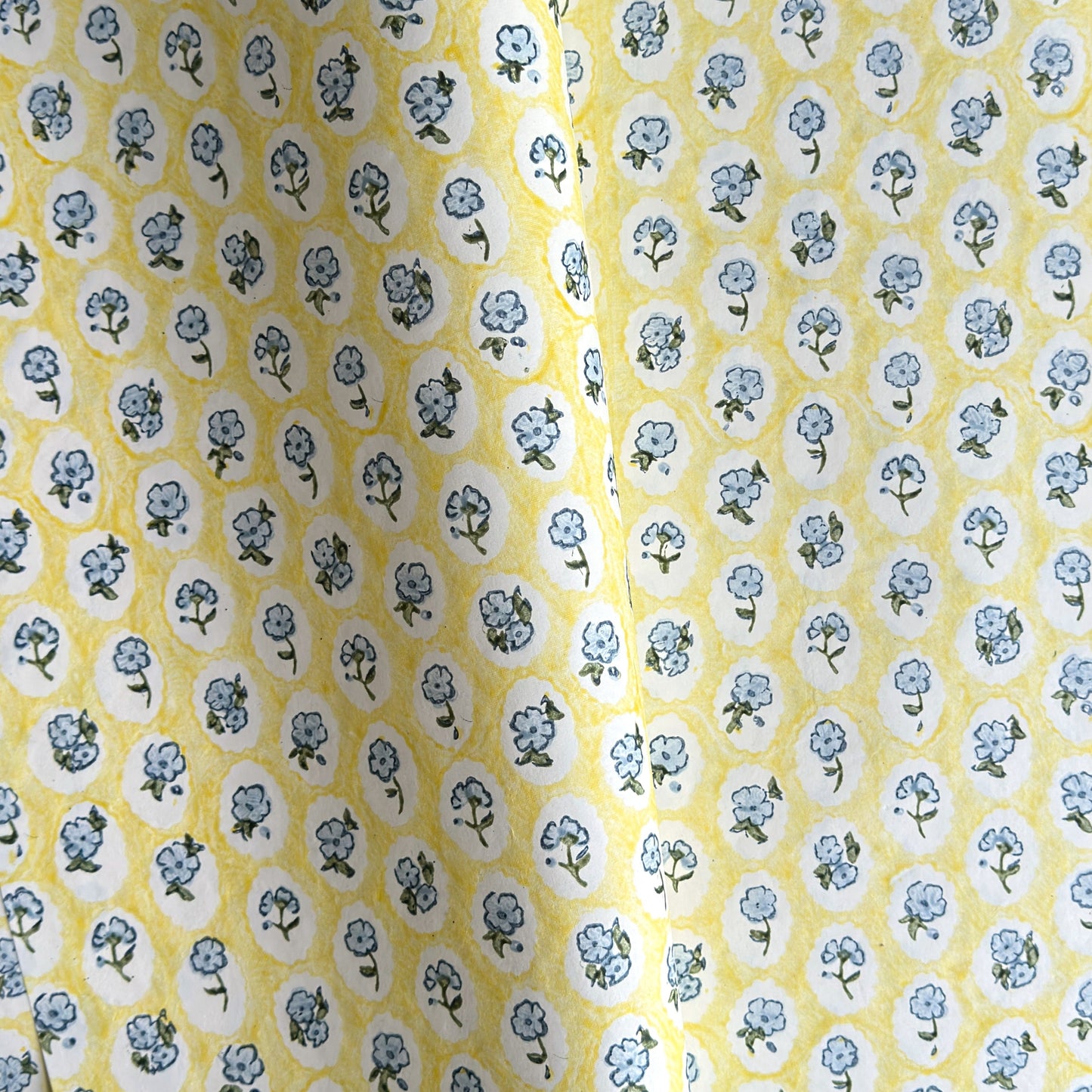 a sheet of hand block printed wrapping paper with a ditsy floral pattern of blue flowers in white ovals with a soft yellow backdrop  