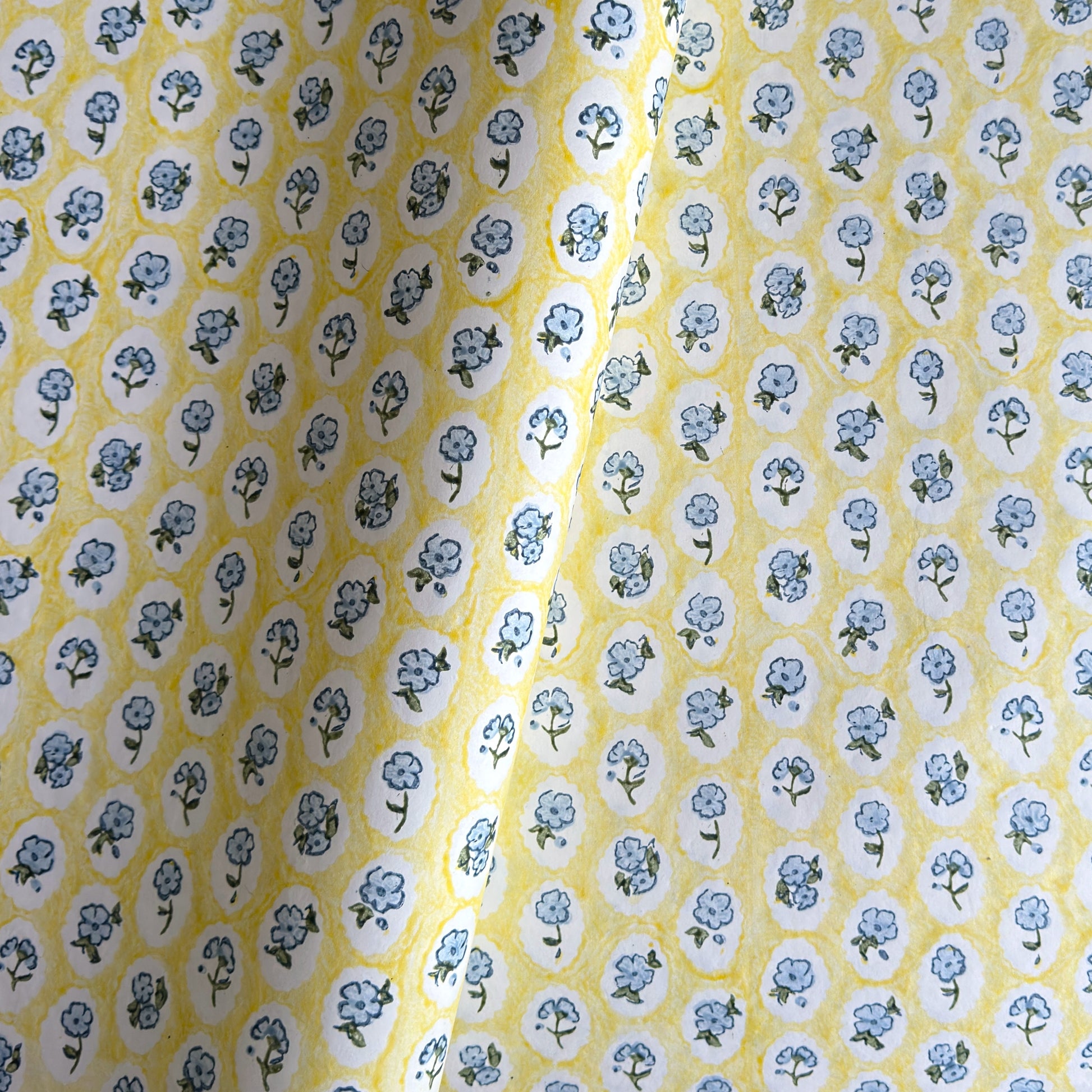 a sheet of hand block printed wrapping paper with a ditsy floral pattern of blue flowers in white ovals with a soft yellow backdrop  