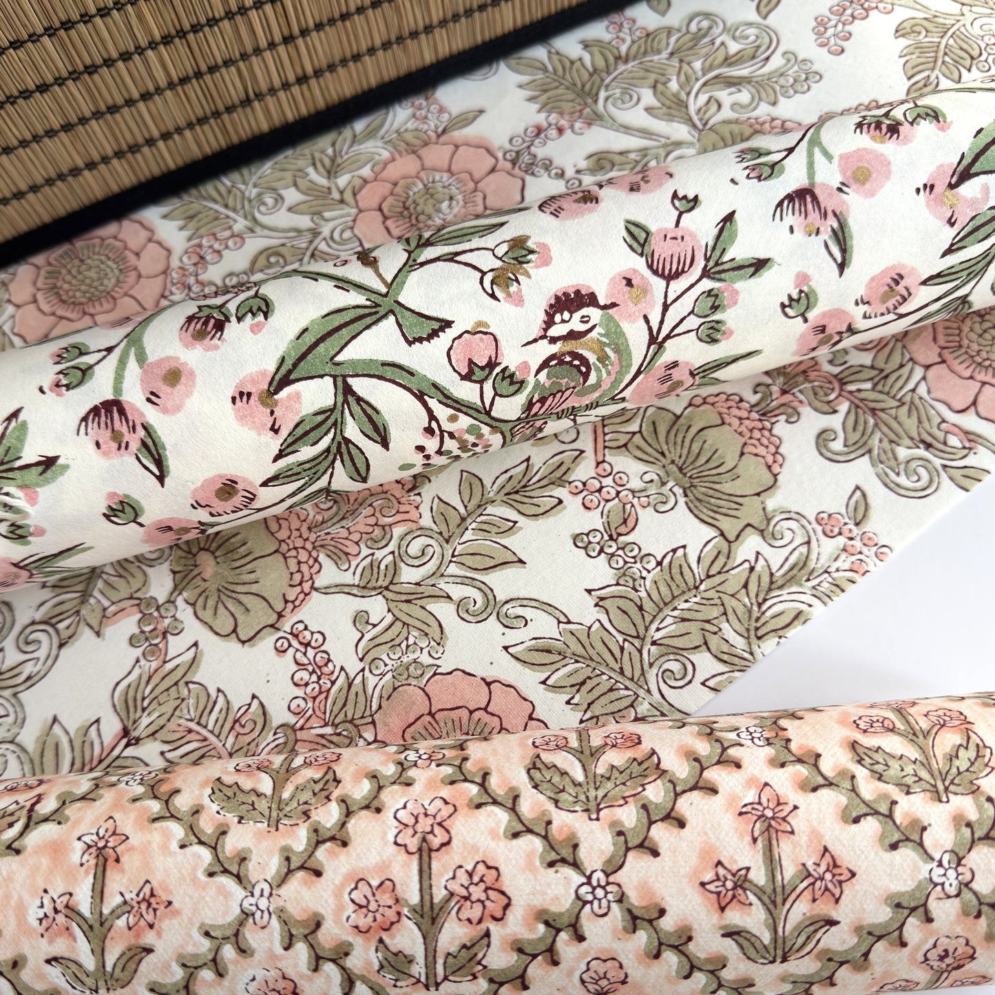 a sheet of hand-block printed wrapping paper with a pattern of a songbird amongst flowering branches in blush pink, soft green and gold. Pictured rolled with other designs