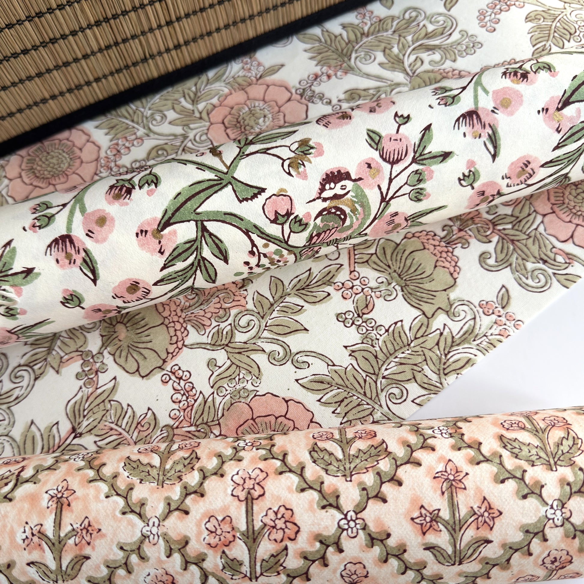 a sheet of hand-block printed wrapping paper with a pattern of a songbird amongst flowering branches in blush pink, soft green and gold. Pictured rolled with other designs
