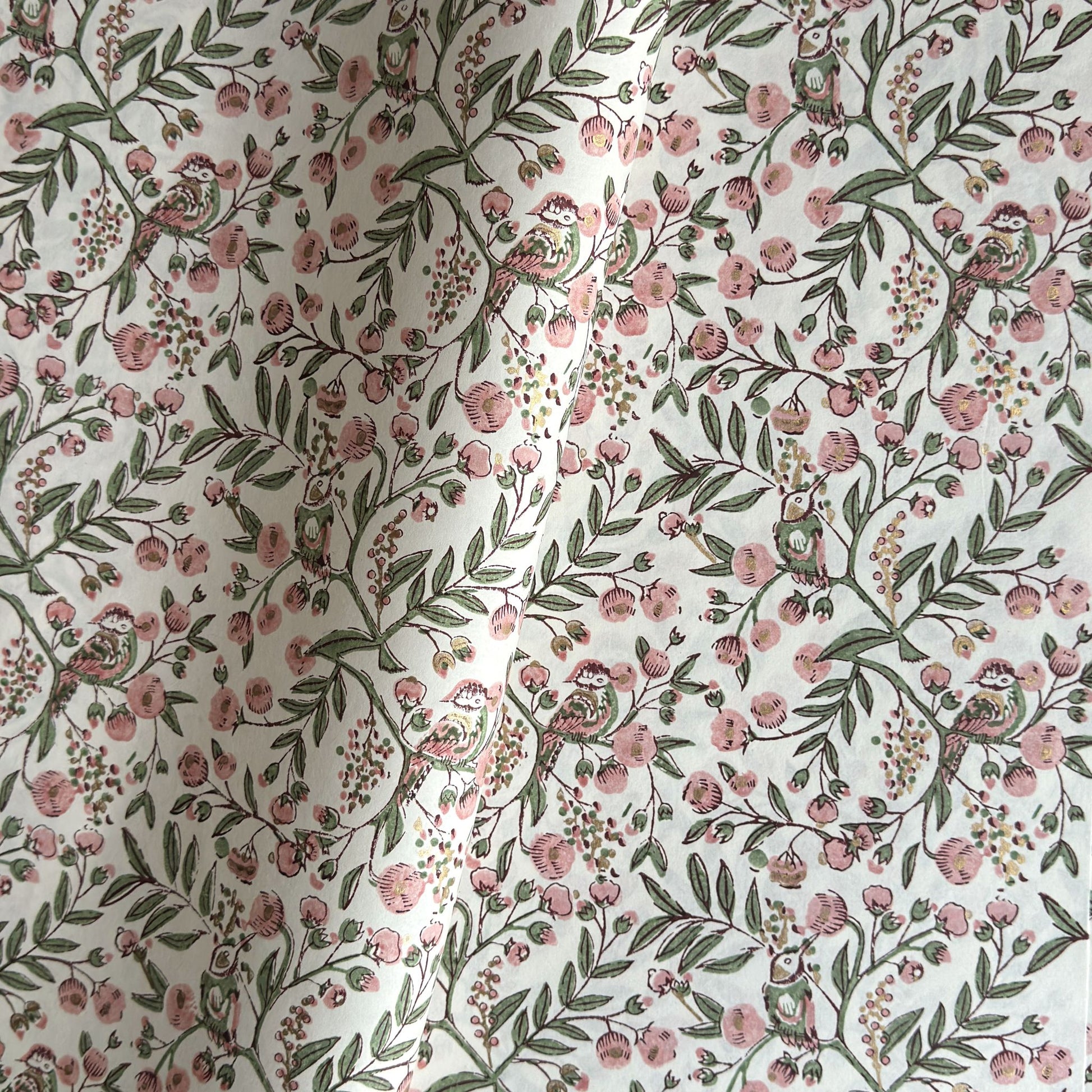 a sheet of hand-block printed wrapping paper with a pattern of a songbird amongst flowering branches in blush pink, soft green and gold