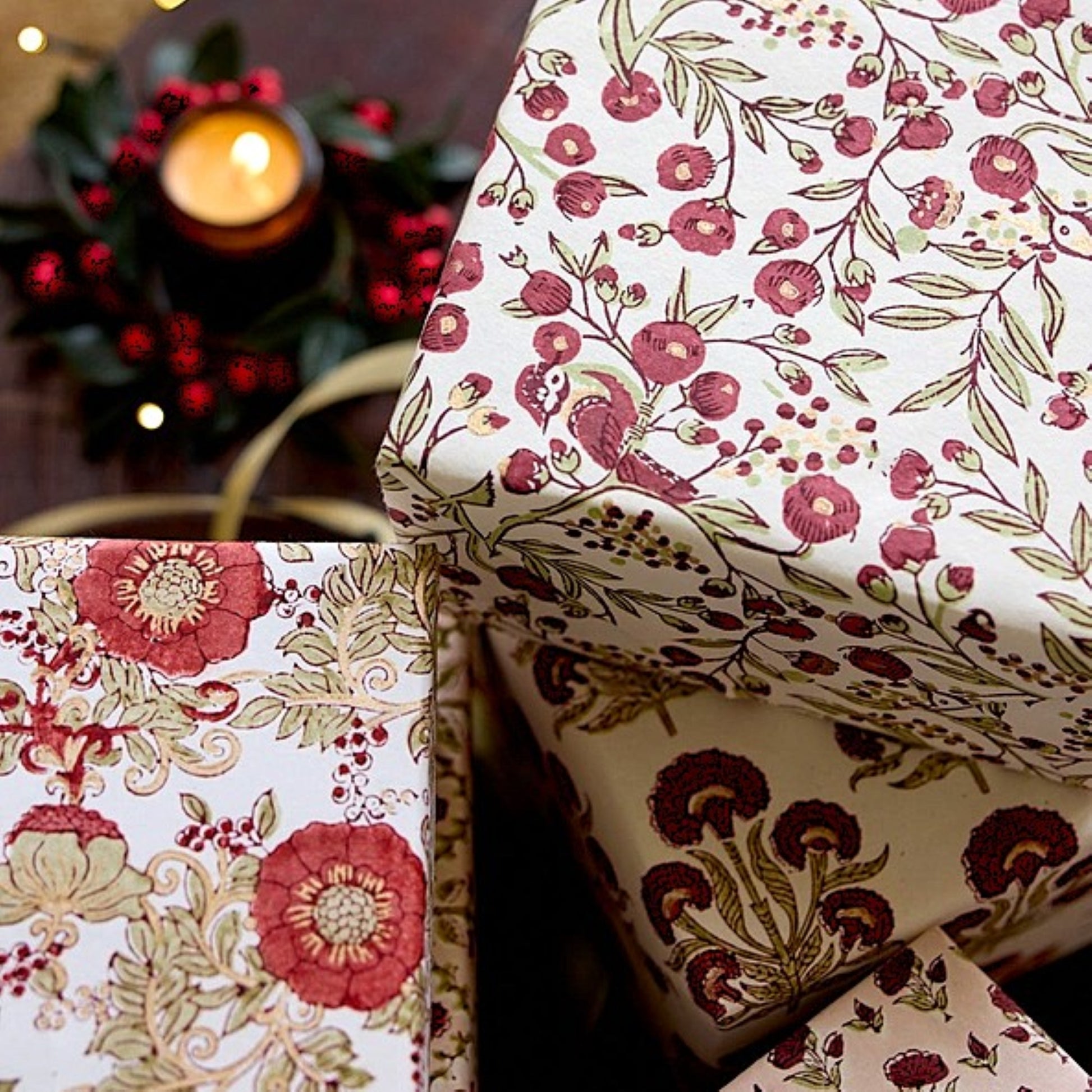 Three gift boxes wrapped in matching floral paper with a vintage botanical pattern, styled with a warm lit candle and red berries in the background.