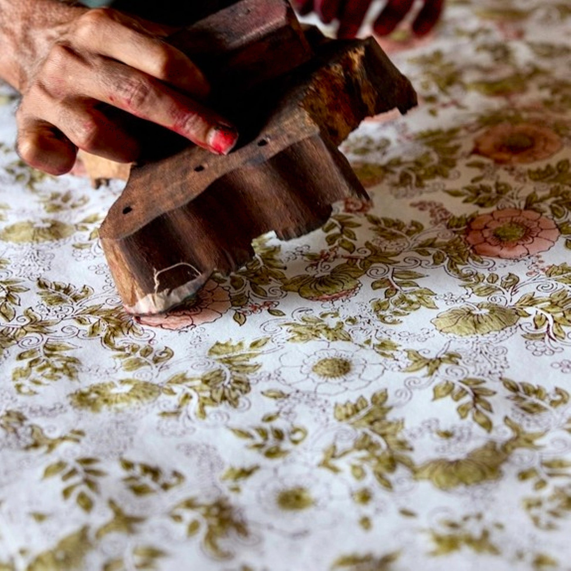 A close-up of a person's hands holding a dark wooden block and hand block printing onto cotton paper a wild rose repeat pattern design.