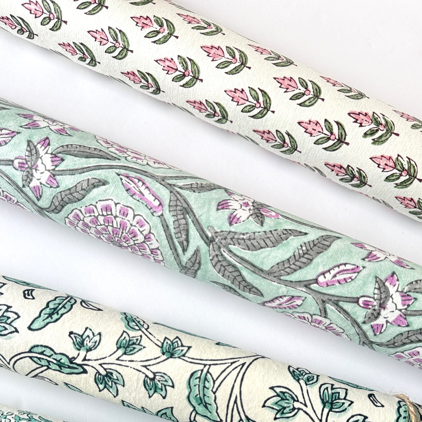 hand block printed paper by Paper Mirchi.  Repeat design of a stylised flower in blush pink and green on a white background. Pictured rolled alongside other designs